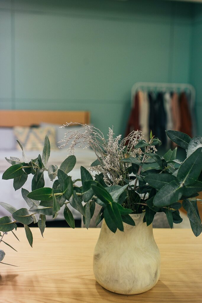 Interior of light room with green eucalyptus in vase placed on wooden surface near bed and wardrobe