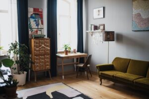 Comfortable couch put on wooden floor covered with rug and potted plants in apartment with big windows and dark curtains in sunlight